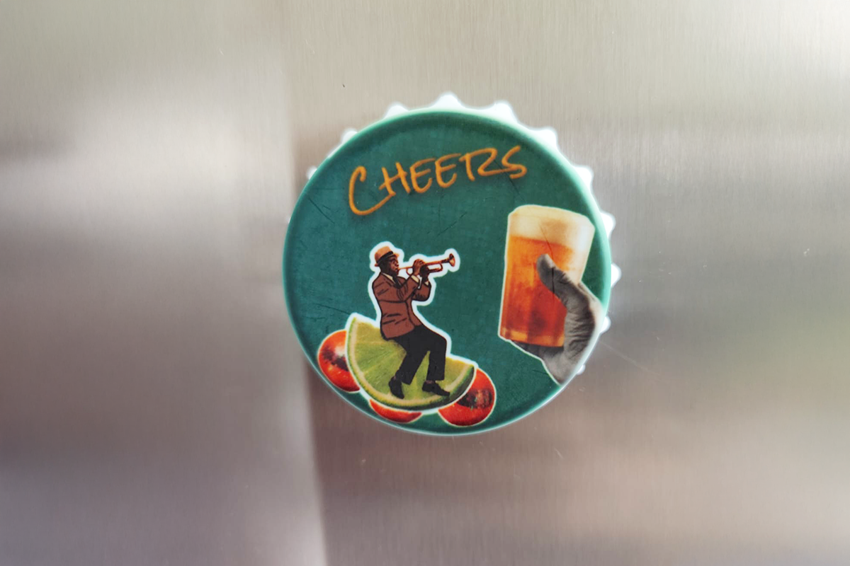 Imã abridor Cheers com arte exclusiva, mostrando um músico de trompete e um brinde com cerveja sobre fundo verde, fixado na porta de uma geladeira.