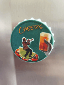 Imagem do imã abridor personalizado com design Cheers, mostrando um músico tocando trompete e um copo de cerveja sobre fundo verde, fixado na porta de uma geladeira.